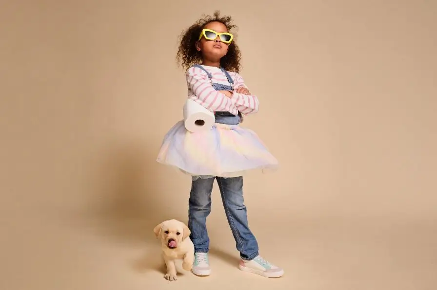 Little girl wearing sunglasses while holding a toilet roll and standing with a Labrador puppy